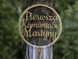Drewniany łapacz snów z napisem "Pierwsza Komunia Święta Martyny" ozdobiony wstążkami w stonowanych kolorach, powieszony na drzewie, w tle las.