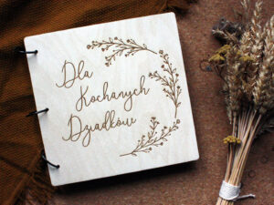 Drewniany album na zdjęcia z napisem na okładce "Dla kochanych Dziadków" leżący na musztardowym pledzie w towarzystwie suszonych kwiatów.