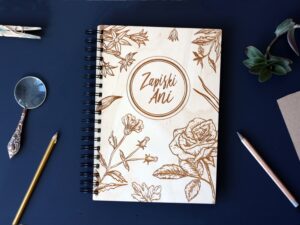 Zdjęcie przedstawia drewniany notes personalizowany "Zapiski Ani" leżący na blacie w towarzystwie lupy i przyborów do pisania.